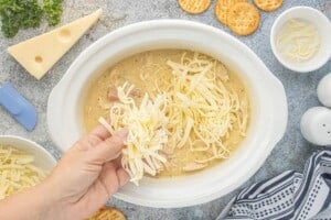 Shredded Swiss and mozzarella cheese being added to the cooked chicken and ham mixture in the slow cooker.
