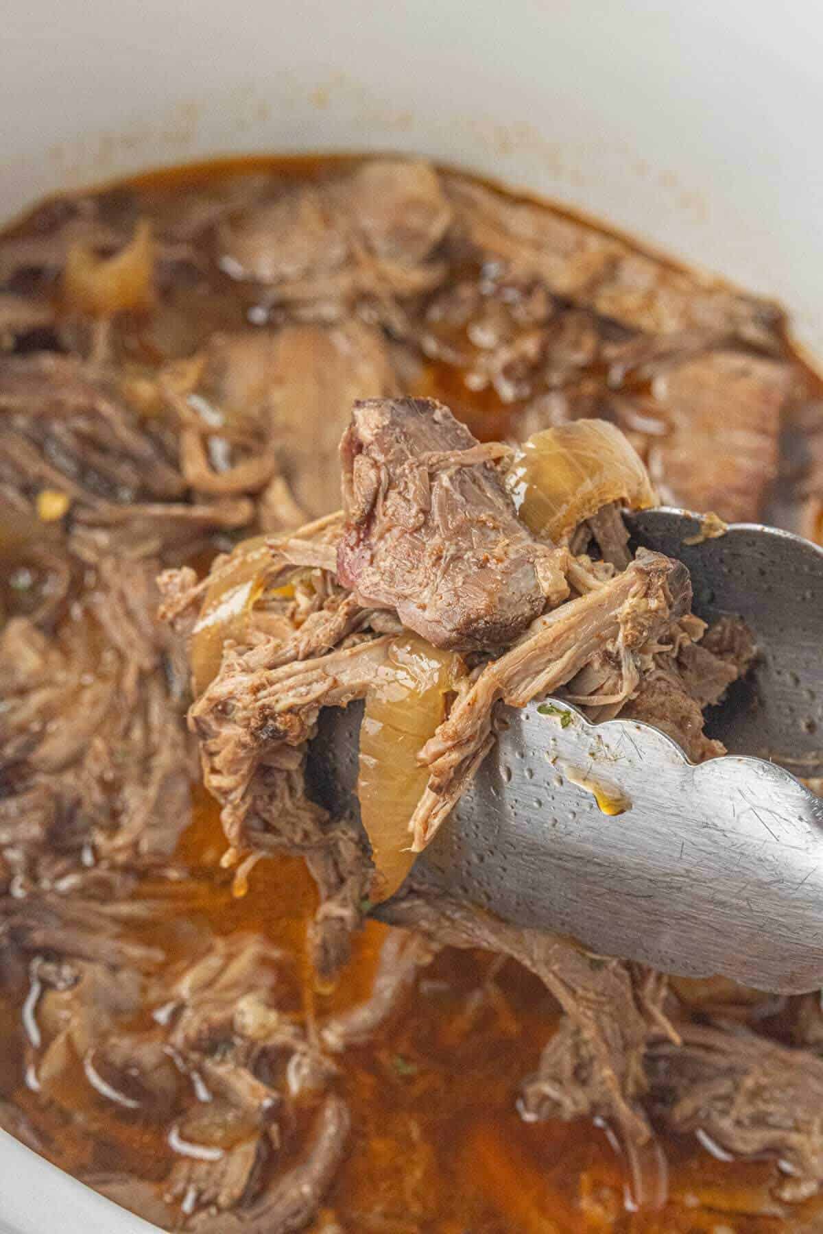 Tender shredded beef and cooked onions lifted from the slow cooker with tongs over the flavorful cooking juices.