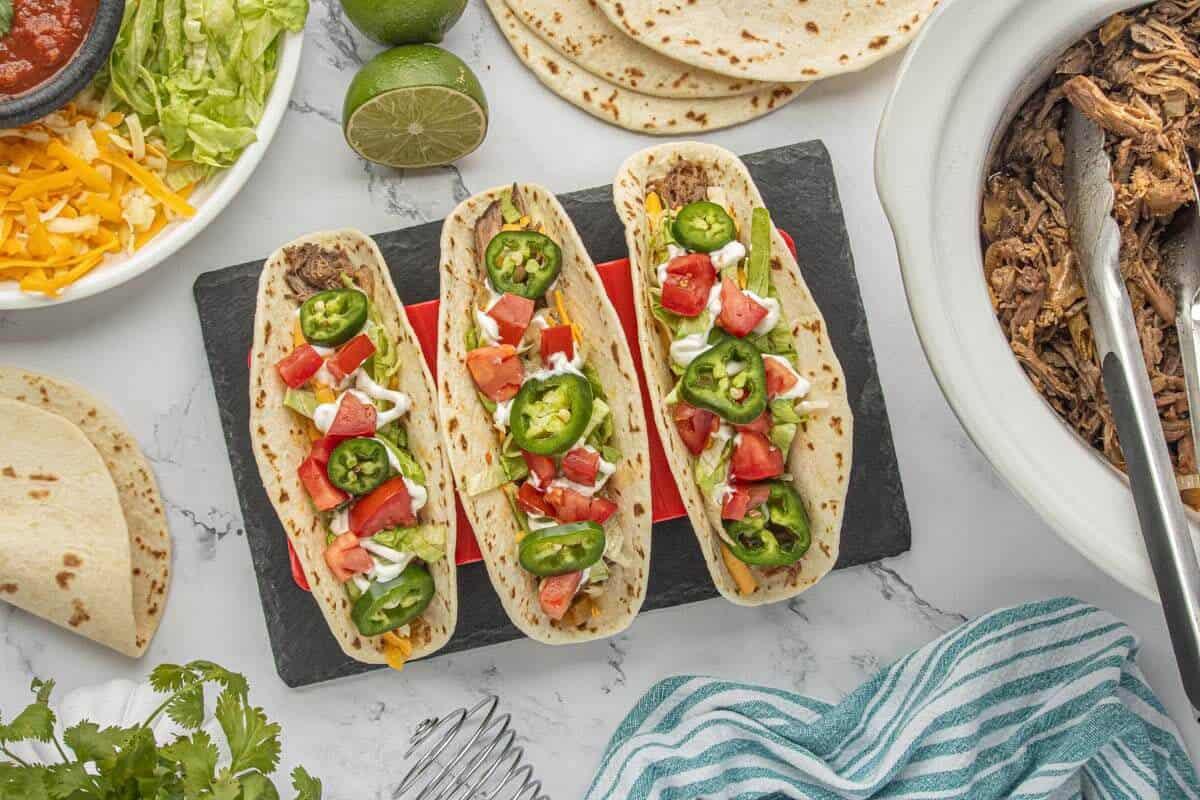 Crockpot shredded beef tacos topped with lettuce, tomatoes, jalapeños and sour cream, served alongside the slow cooker.
