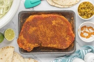 Chuck roast coated with seasoning on a baking sheet with tortillas, green chiles and limes nearby.
