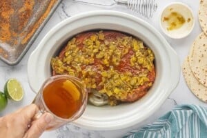 Seasoned chuck roast topped with green chiles in a slow cooker as beef broth is poured over the meat.