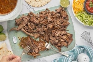Slow-cooked chuck roast being shredded with two forks on a cutting board beside taco toppings.