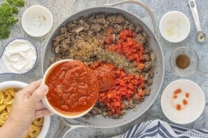Marinara sauce being added to seasoned ground beef, onions, diced tomatoes, and Italian seasoning in a large skillet.