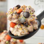 Close-up of crockpot candy peanuts with orange and black M&M's and colorful sprinkles served on a black spoon.