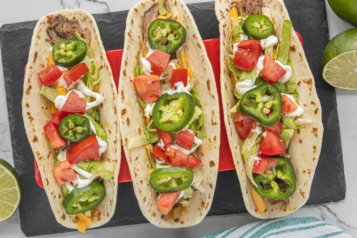 Three shredded beef tacos topped with lettuce, tomatoes, jalape&ntilde;os and sour cream on a black serving board with fresh limes.