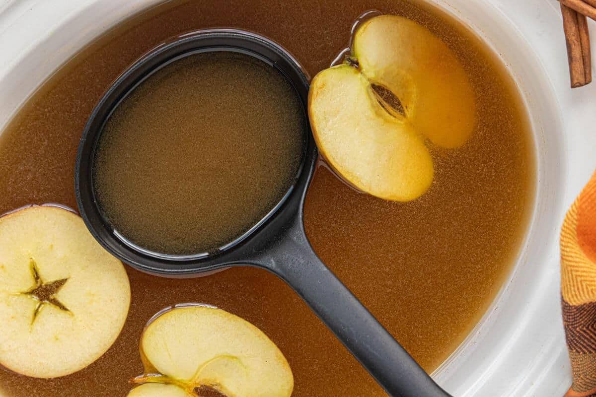 Overhead close-up of Crockpot Caramel Apple Cider with fresh apple slices floating in the warm, spiced cider and a black ladle ready for serving.