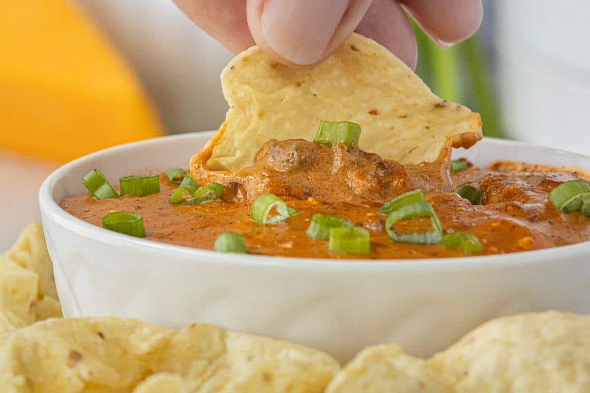 Close-up of a tortilla chip loaded with thick, creamy chili and melted cheese, lifted from a white serving bowl topped with green onions.