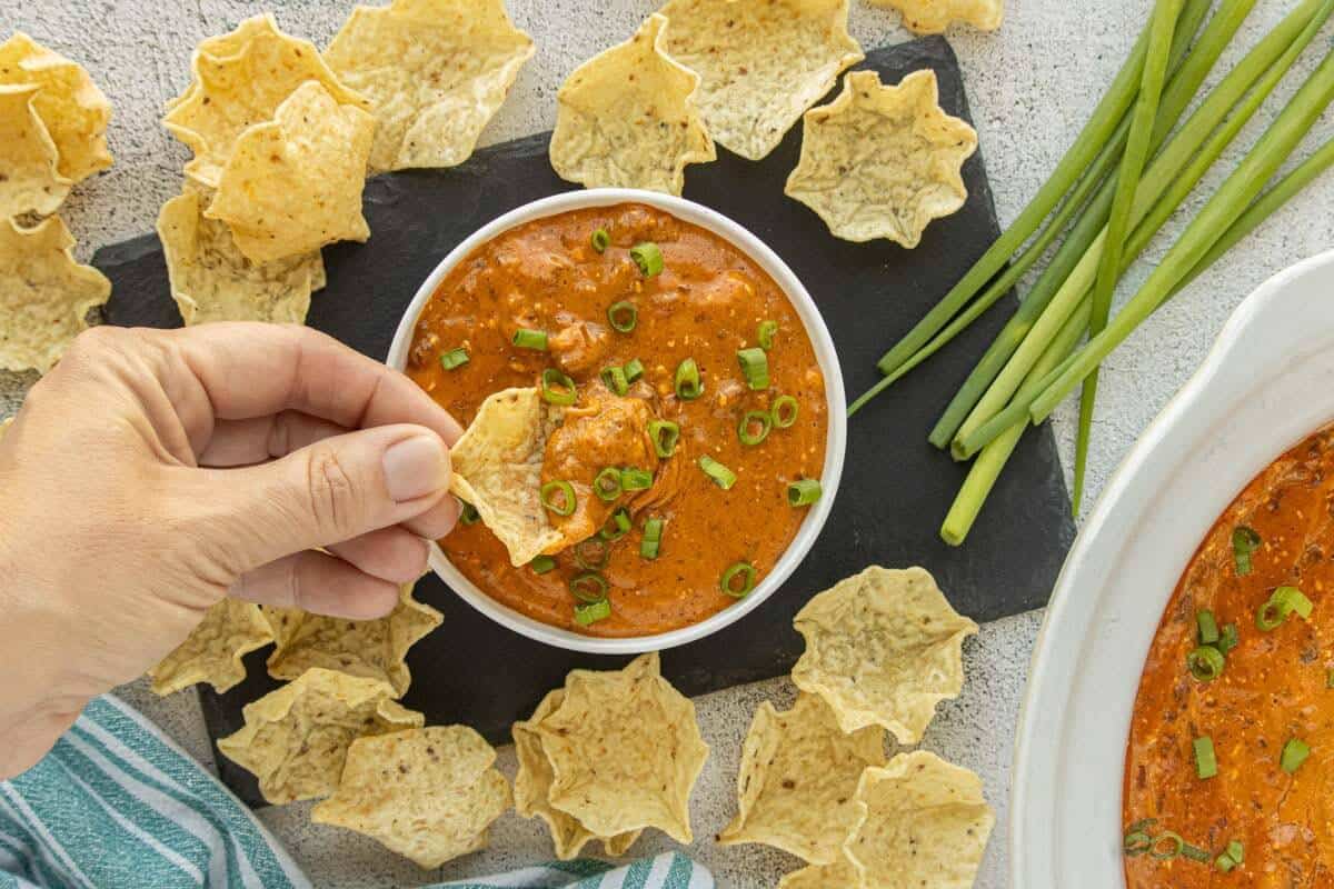 Overhead view of a tortilla chip scooping up cheesy chili dip, surrounded by tortilla chips, fresh green onions, and a slow cooker.