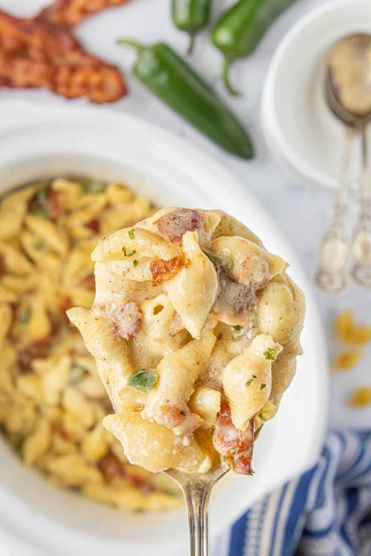 Close-up of a spoonful of Crockpot Bacon Jalapeño Mac and Cheese with creamy pasta shells, crispy bacon, and diced jalapeños, held above a white crockpot.