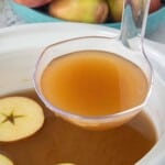 Close-up of a ladle filled with warm caramel apple cider being scooped from a white slow cooker, with a floating apple slice and fresh apples in the background.