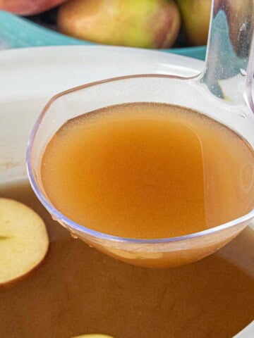 Close-up of a ladle filled with warm caramel apple cider being scooped from a white slow cooker, with a floating apple slice and fresh apples in the background.
