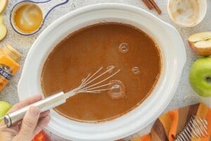 Caramel apple cider being stirred with a whisk in a white crockpot, surrounded by fresh apples, cinnamon sticks, and caramel sauce.