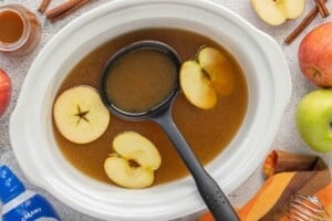 Homemade caramel apple cider in a white slow cooker with floating apple slices and a black ladle, ready to serve.