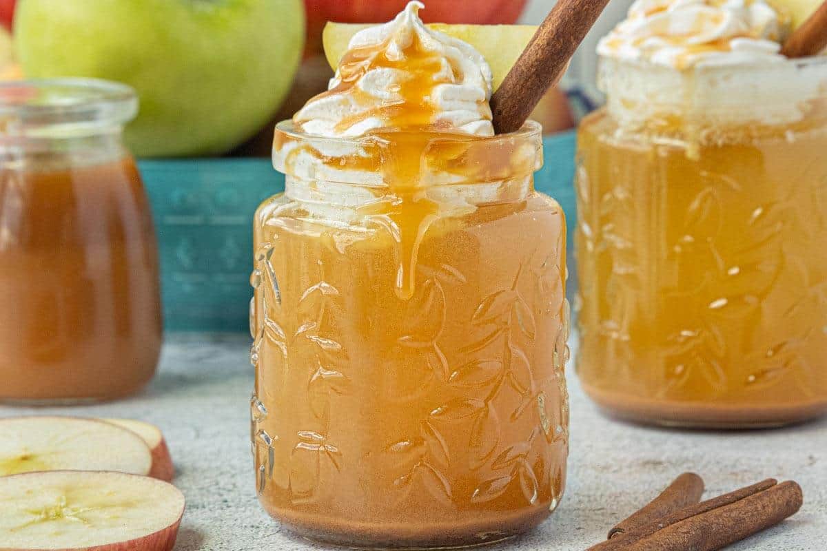 Crockpot caramel apple cider served in a glass mason jar, topped with whipped cream, caramel drizzle, and a cinnamon stick, with fresh apples and another mug of cider in the background.