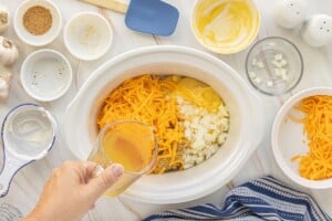 Chicken broth being poured into a slow cooker with rice, cheddar cheese, diced onion.