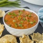 Creamy Crockpot Chili Cheese Dip garnished with sliced green onions in a white bowl, surrounded by crispy tortilla chips for dipping.