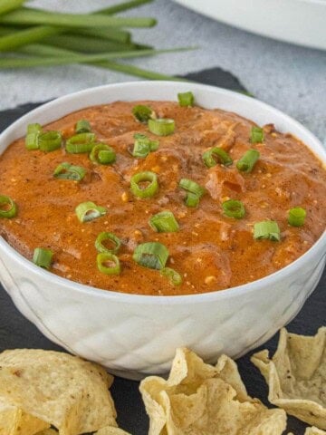 Creamy Crockpot Chili Cheese Dip garnished with sliced green onions in a white bowl, surrounded by crispy tortilla chips for dipping.