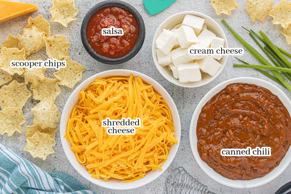 Overhead view of ingredients for Crockpot chili cheese dip, including canned chili, salsa, cubed cream cheese, freshly shredded cheddar, and tortilla scoops.