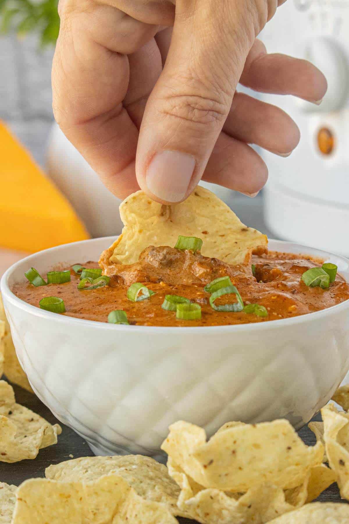 Hand dipping a crispy tortilla chip into a bowl of creamy chili cheese dip garnished with sliced green onions.