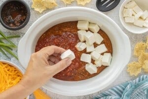 Hand adding cubes of cream cheese to chili in a white slow cooker to make Crockpot Chili Cheese Dip.