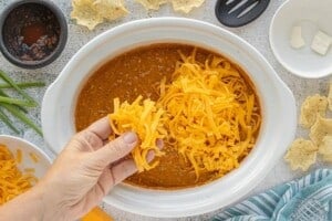 Hand sprinkling freshly shredded cheddar cheese over creamy chili dip in a white crockpot before melting.