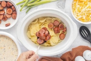 Browned sausage slices being spooned over a layer of dried scalloped potatoes in a white slow cooker.
