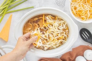 Freshly shredded cheddar and Monterey Jack cheese being sprinkled over the cooked sausage and potato casserole in a crockpot.