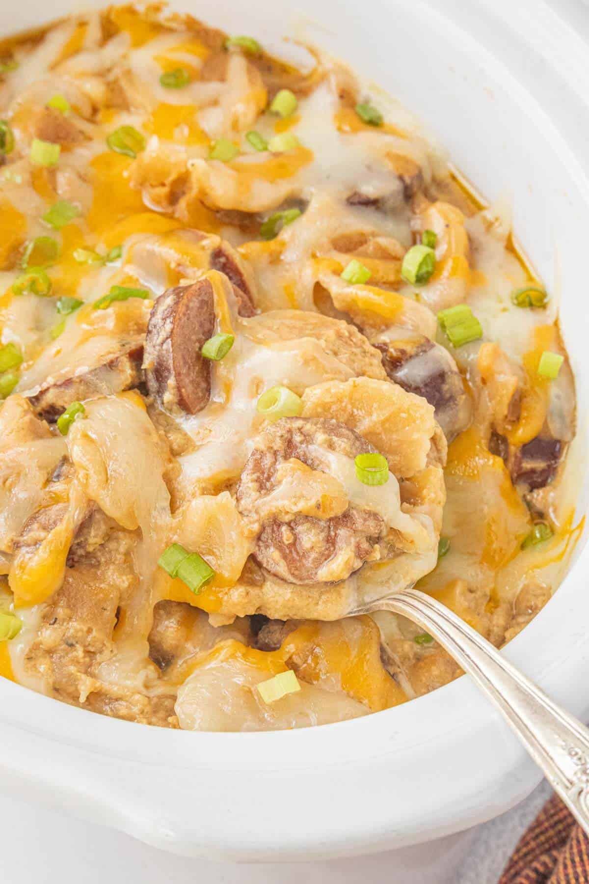Creamy scalloped potatoes and browned kielbasa topped with melted cheddar cheese and chopped green onions in a white slow cooker, with a serving spoon ready to scoop out a spoonful.