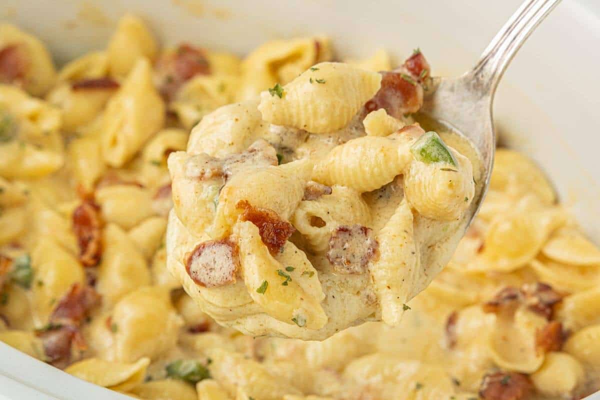 Heaping spoonful of Crockpot Bacon Jalape&ntilde;o Mac and Cheese lifted above a dish filled with cheesy pasta shells, crumbled bacon, and green jalape&ntilde;os.