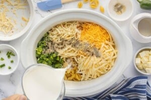 Pouring milk into a Crockpot filled with uncooked pasta shells, shredded cheese, diced jalapeños, and seasonings for Bacon Jalapeño Mac and Cheese.