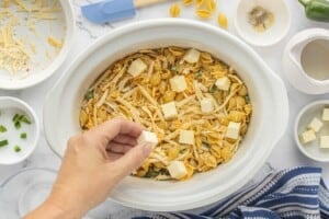 Adding cubes of butter to the uncooked pasta, shredded cheese, and jalapeño mixture in a slow cooker.