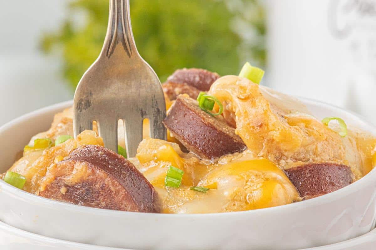 Close-up of browned sausage slices and cheesy potatoes in a white bowl, garnished with chopped green onions and a fork ready for a bite.