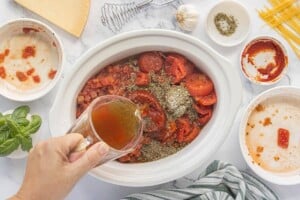 Beef broth being poured into a slow cooker filled with tomatoes, browned meat, and Italian seasonings.