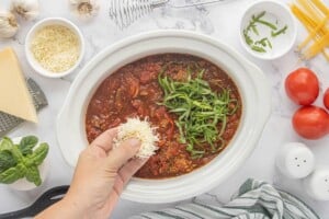 Freshly grated Parmesan cheese being added to a rich, chunky tomato meat sauce topped with sliced fresh basil in a slow cooker.