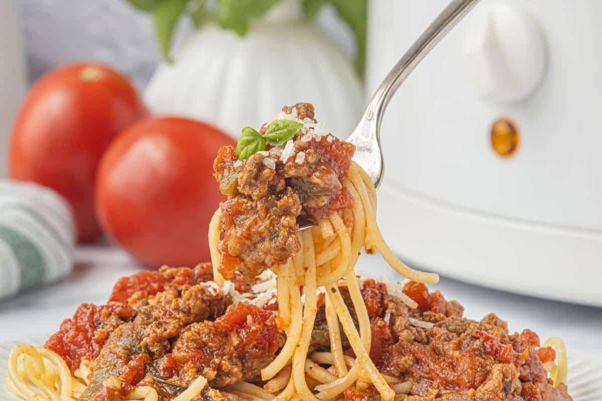 Fork lifting spaghetti with hearty beef and sausage sauce above a plate of pasta, with fresh tomatoes and a white slow cooker in the background.
