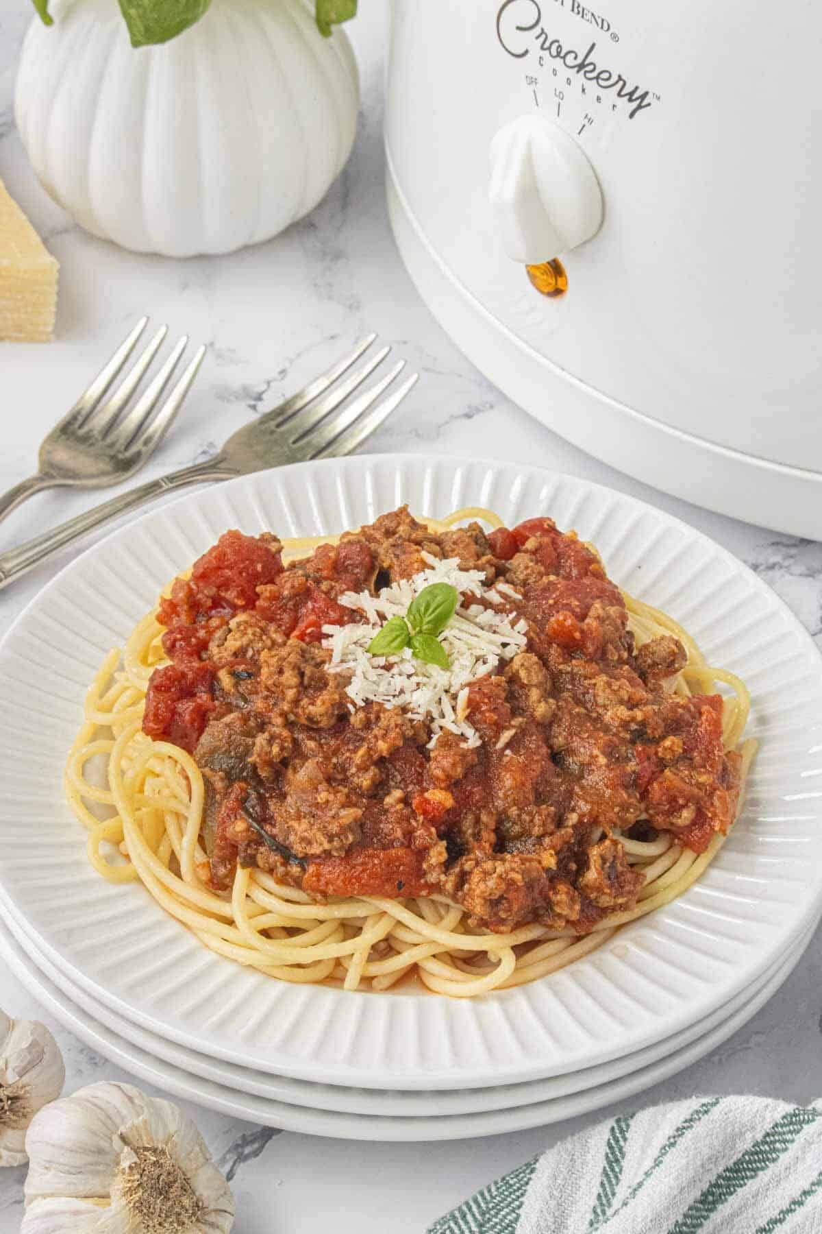 Plate of spaghetti topped with hearty tomato meat sauce, shredded Parmesan cheese, and fresh basil, served beside a white slow cooker.