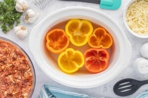 Five hollowed-out red, orange, and yellow bell peppers arranged upright in a white crockpot, ready to be stuffed with ground beef and rice filling.