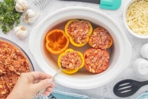 Ground beef and rice filling being spooned into colorful bell peppers arranged in a white slow cooker before cooking.
