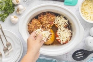 Shredded mozzarella cheese being sprinkled over cooked stuffed bell peppers filled with ground beef, rice, and tomato sauce in a crockpot.