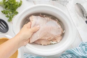 Raw boneless turkey breast being placed over sliced onions in a white slow cooker.