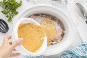 Creamy seasoned turkey gravy being poured over a raw turkey breast in a white crockpot.