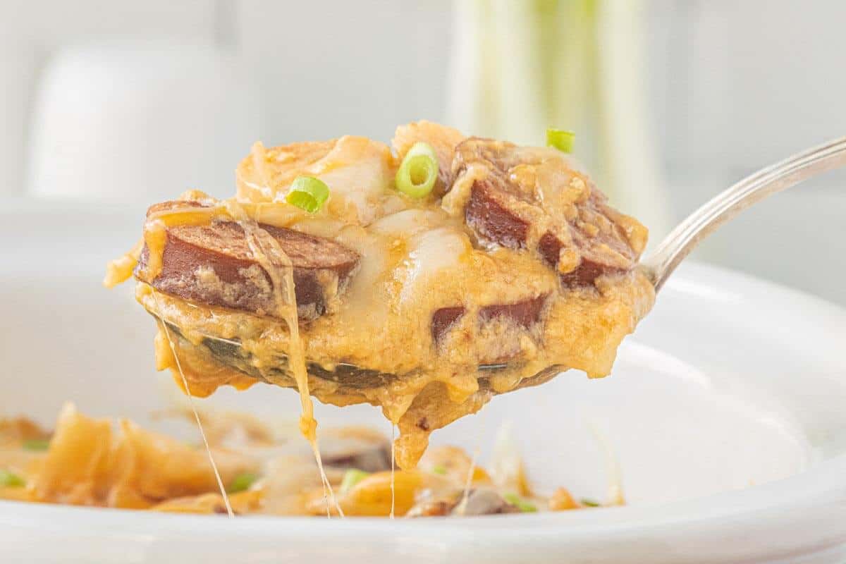A spoonful of Crockpot kielbasa and potatoes lifted from a slow cooker, with strings of melted cheese stretching toward the dish.