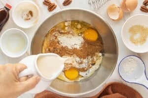 Evaporated milk being poured into a mixing bowl with pumpkin puree, eggs, brown sugar, and spices.