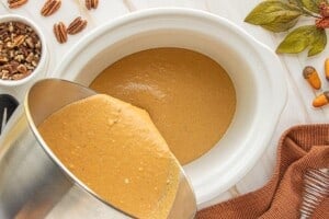 Pumpkin pie pudding batter being poured from a mixing bowl into a white slow cooker.