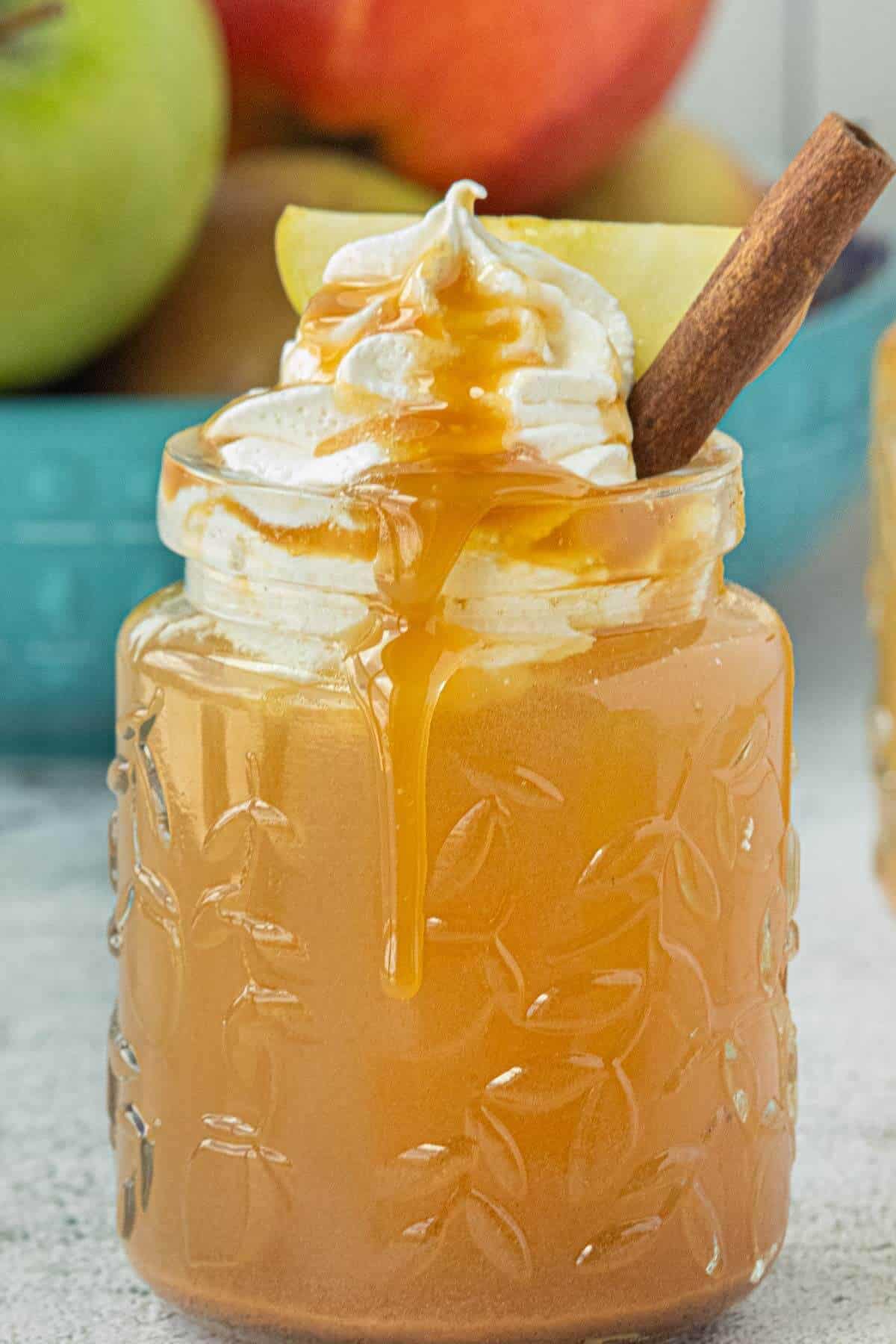 Slow Cooker Caramel Apple Cider in a glass mason jar topped with whipped cream, caramel drizzle, an apple slice, and a cinnamon stick, with caramel dripping down the side.
