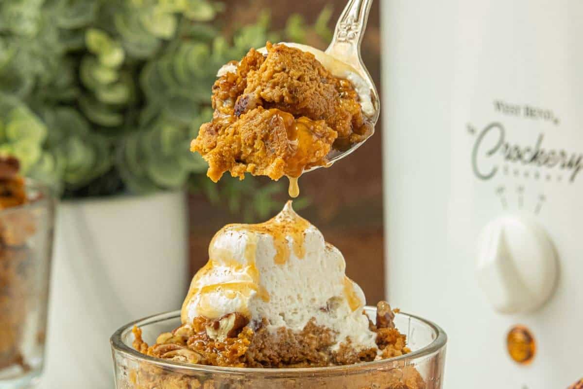 Spoonful of pumpkin pie pudding drizzled with caramel sauce above a glass dessert cup topped with whipped cream.