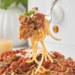 Close-up of a forkful of spaghetti coated in chunky tomato meat sauce, topped with grated Parmesan cheese and fresh basil.