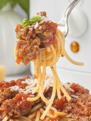 Close-up of a forkful of spaghetti coated in chunky tomato meat sauce, topped with grated Parmesan cheese and fresh basil.